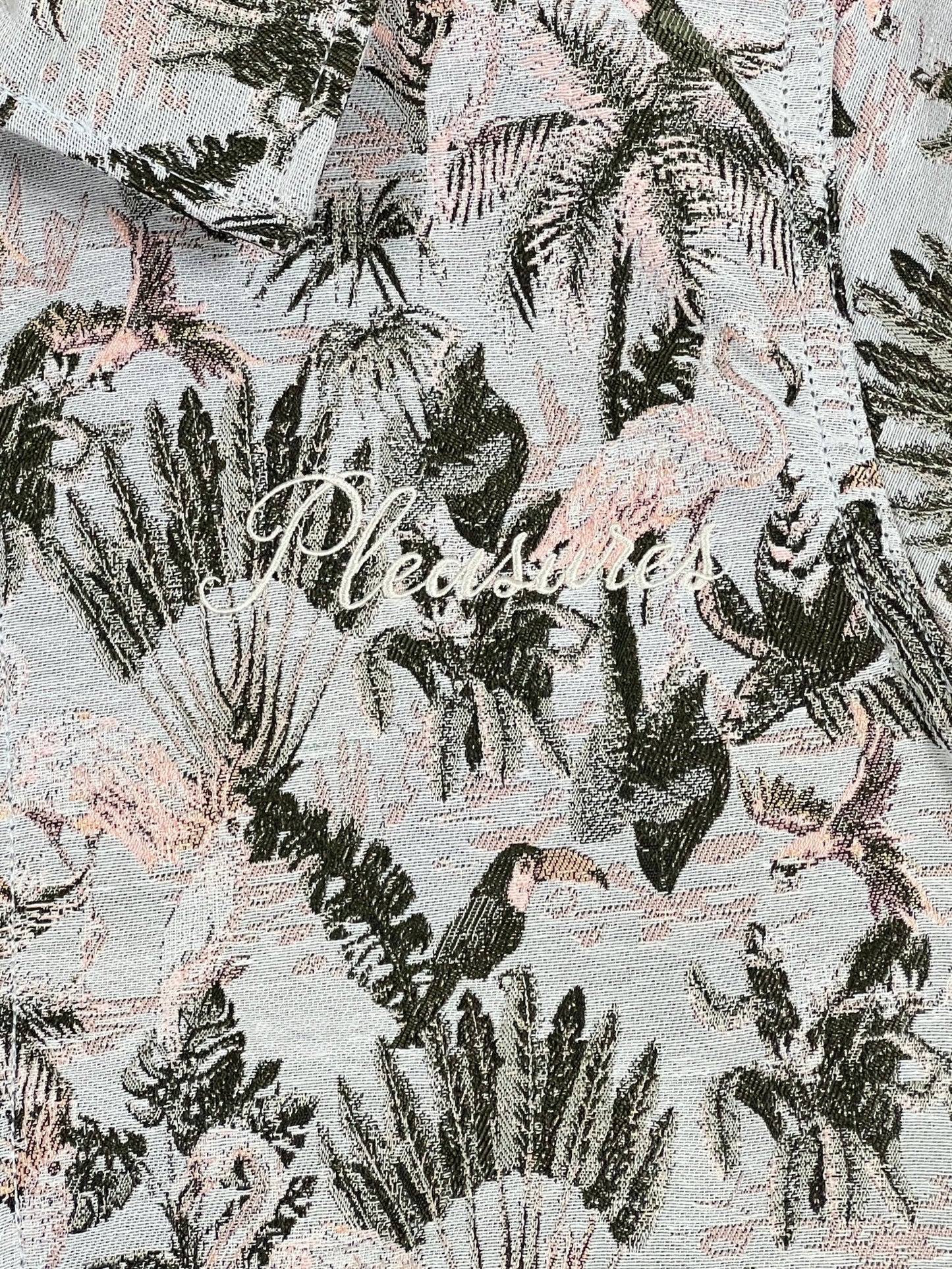 A close up of a PLEASURES PARROT WORK JACKET GREY Hawaiian print shirt with front zip closure.