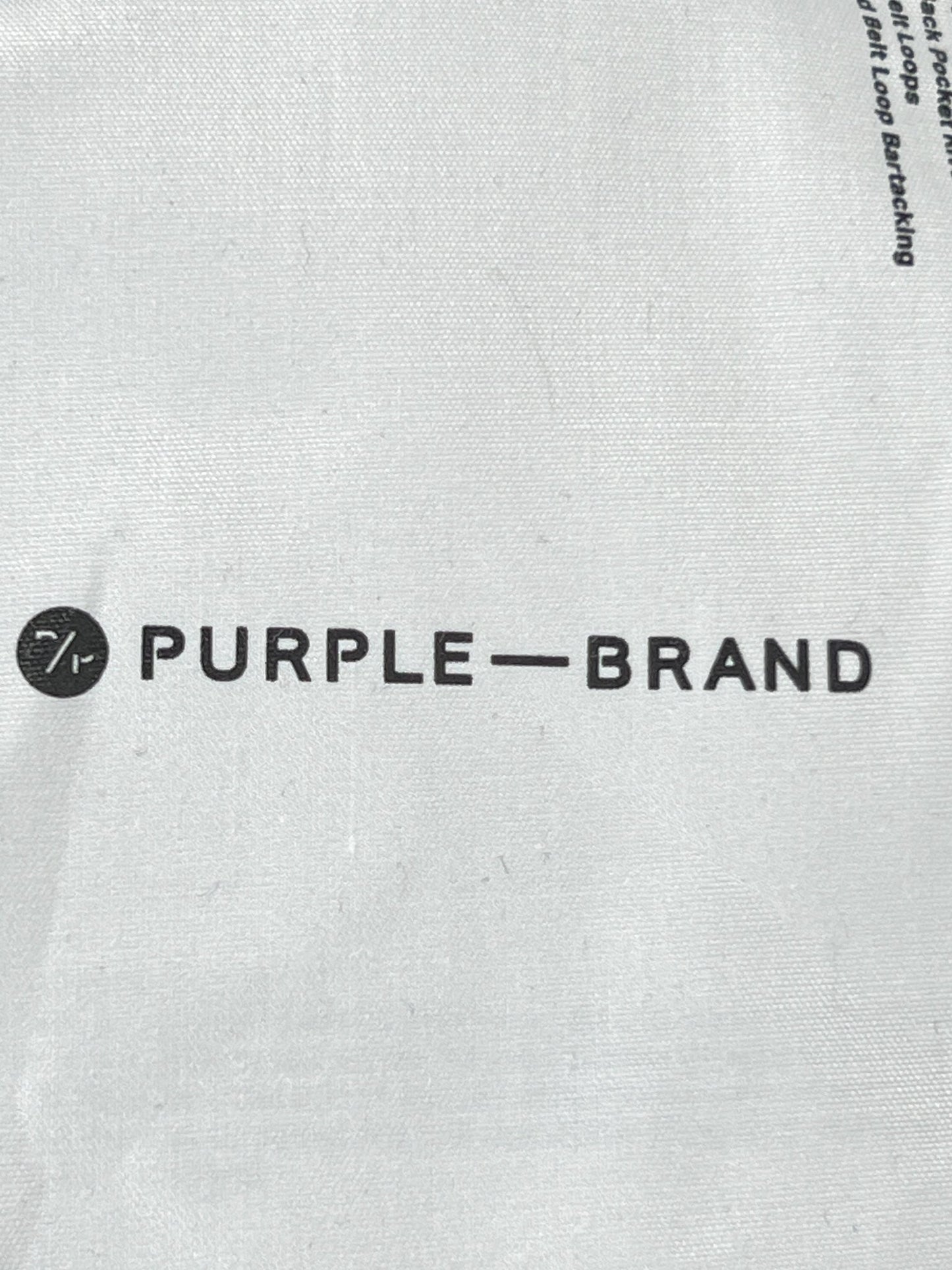 The distressed PURPLE BRAND logo on a white bag featuring the PURPLE BRAND P004-PRFL PATCH REPAIR FLARE MID INDIGO.