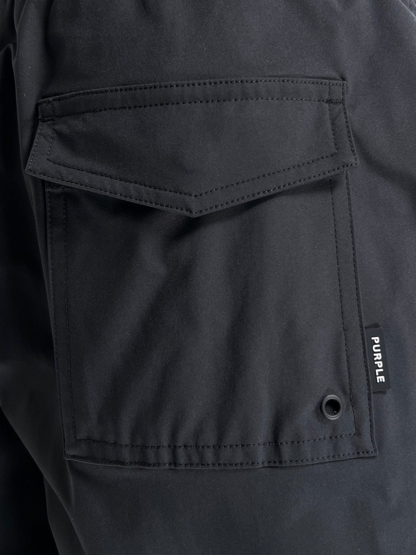 Close-up of a black Polyester swim trunks pocket with a button snap and a brand tag reading "PURPLE BRAND P504-PBUC ALL ROUND SHORT BLACK.
