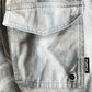 Close-up of a light indigo denim jacket pocket showcasing a bold black label with "PURPLE" in white, crafted by PURPLE BRAND, resembling the P504-PIDD All Round Short Water Print Lt Indigo design.