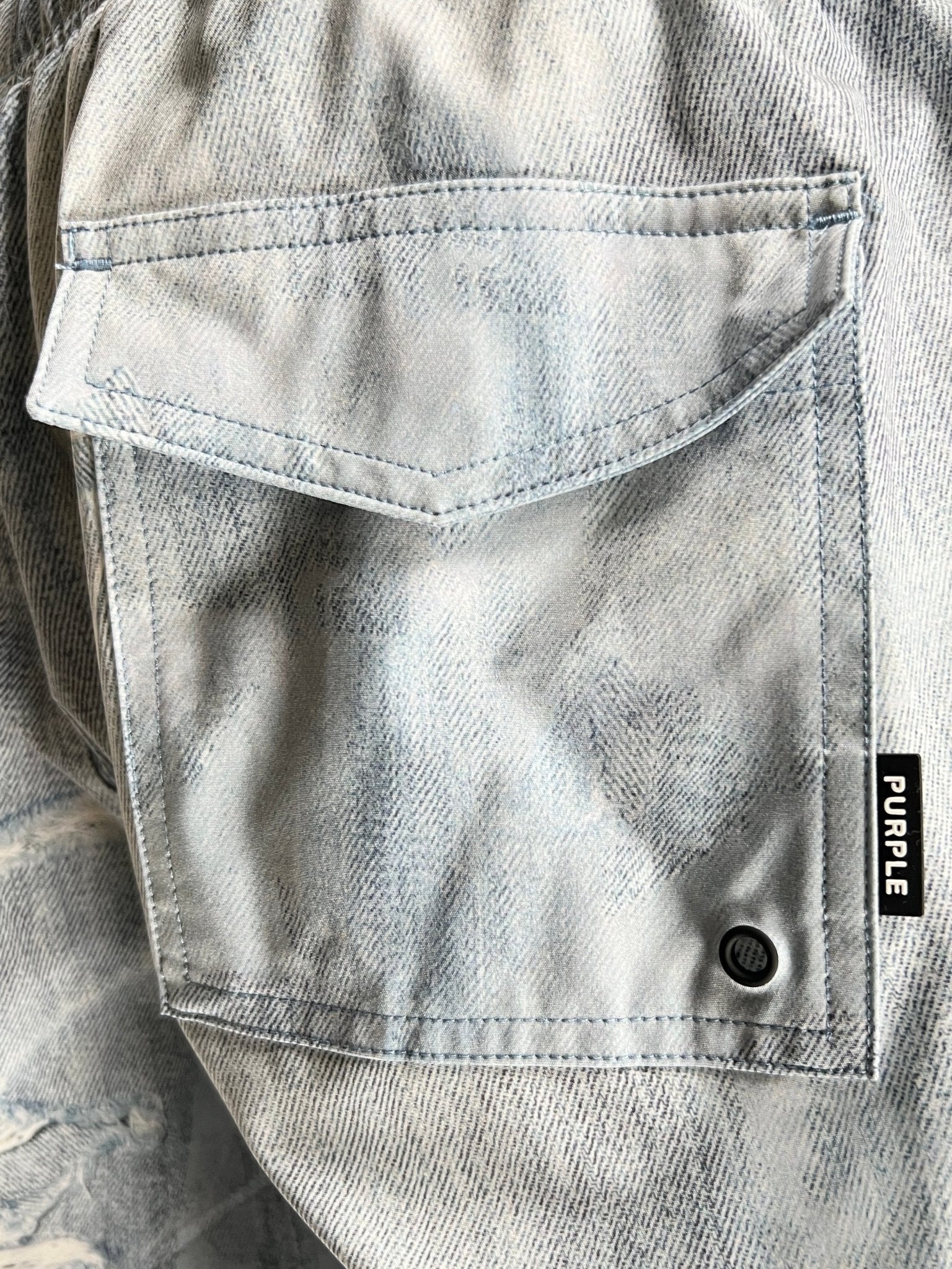 Close-up of a light indigo denim jacket pocket showcasing a bold black label with "PURPLE" in white, crafted by PURPLE BRAND, resembling the P504-PIDD All Round Short Water Print Lt Indigo design.