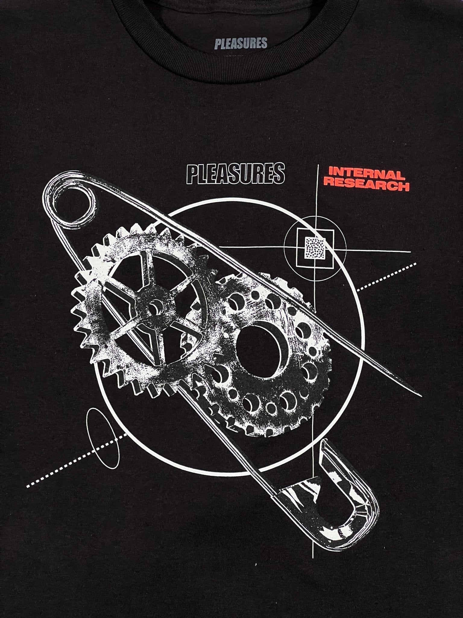 The Research T-Shirt Black/Black by PLEASURES is made from 100% cotton, showcasing white and red abstract gears and circles, with "PLEASURES" and "INTERNAL RESEARCH" elegantly printed on it.